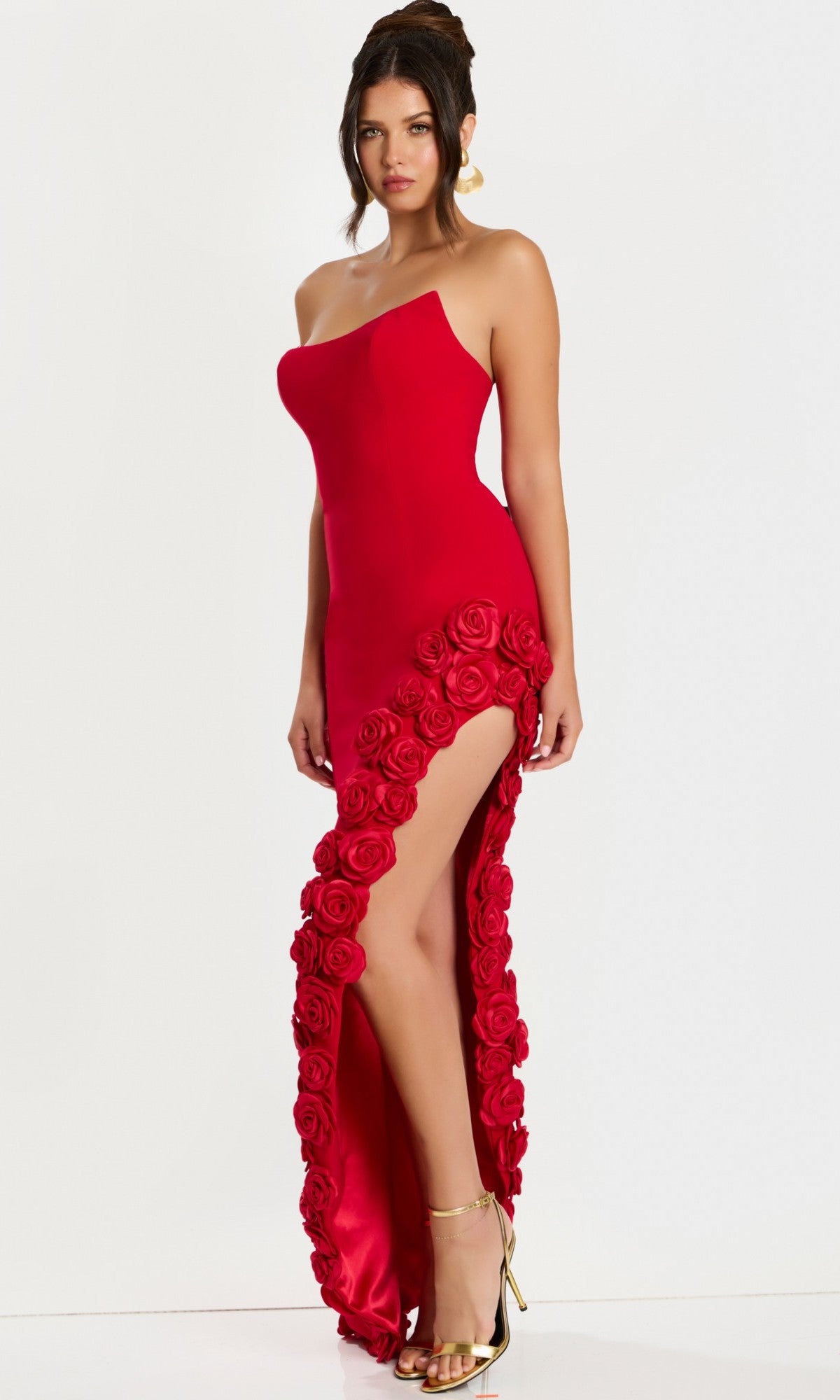 A woman wears the Jovani 47798 long prom dress—a strapless, fitted crepe column with a high slit and 3D floral appliques on the hem—paired with gold heels, standing against a plain white background.